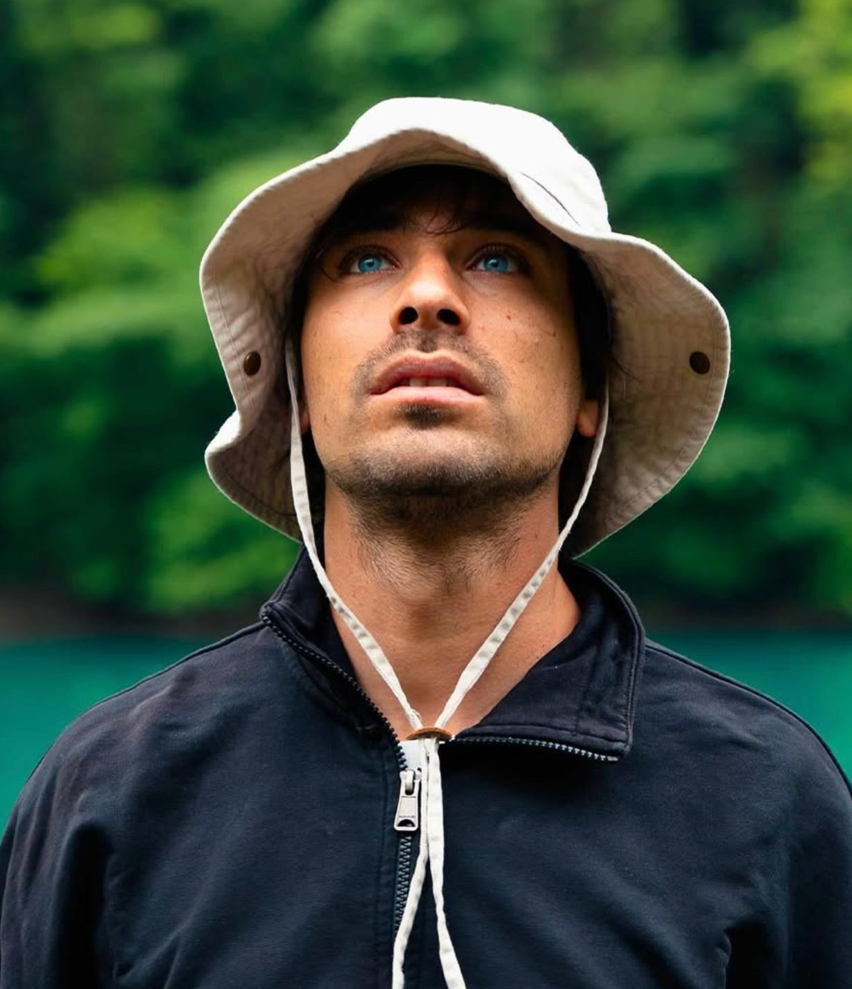 Uomo con occhi azzurri che indossa un bucket hat bianco e una giacca con zip nera guardando verso l'alto con sfondo di fogliame verde.